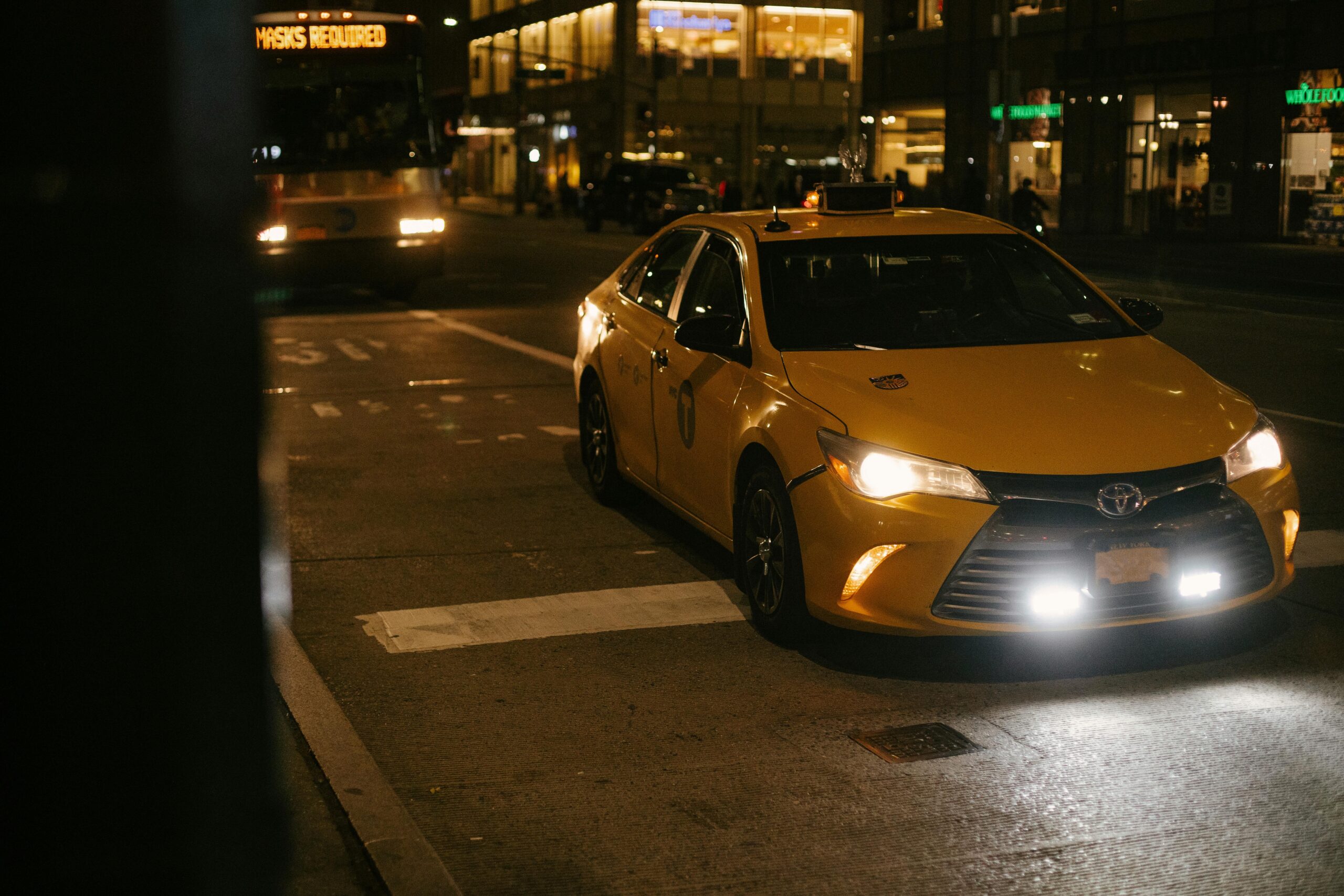 A yellow taxi cab waits at a city street intersection at night, illuminated by headlights.